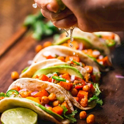 A hand squeezes lime juice over freshly prepared vegetable tacos on a wooden board. The tacos are garnished with chickpeas, avocado, and cilantro.
