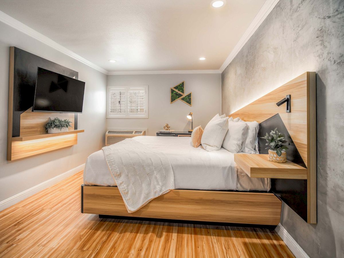 A modern hotel-style bedroom with a wooden platform bed, wall-mounted TV, floating nightstands, and a light gray accent wall.