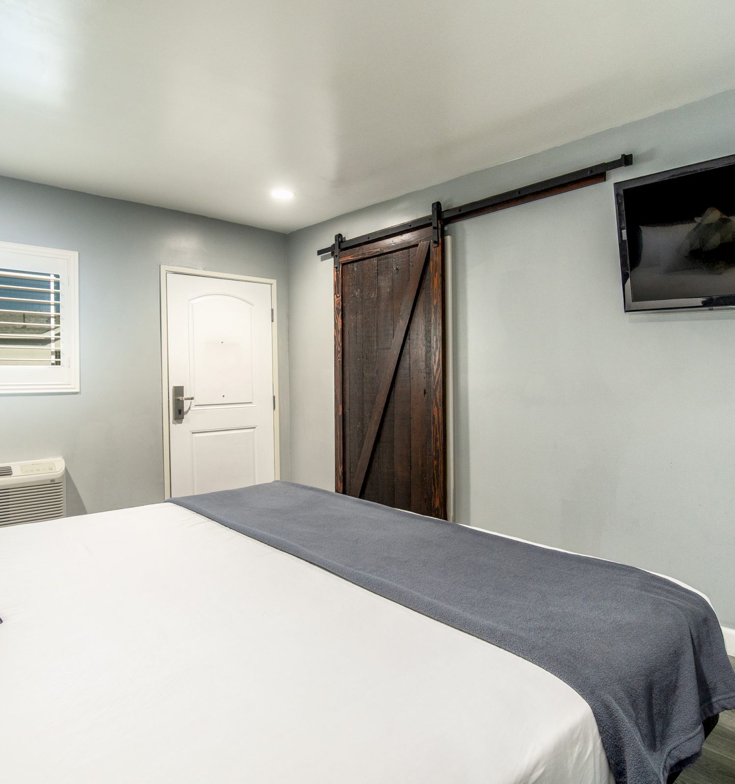 The image shows a modern bedroom with a bed, a sliding barn door, a flat-screen TV, and a window with white shutters. The walls are painted gray.