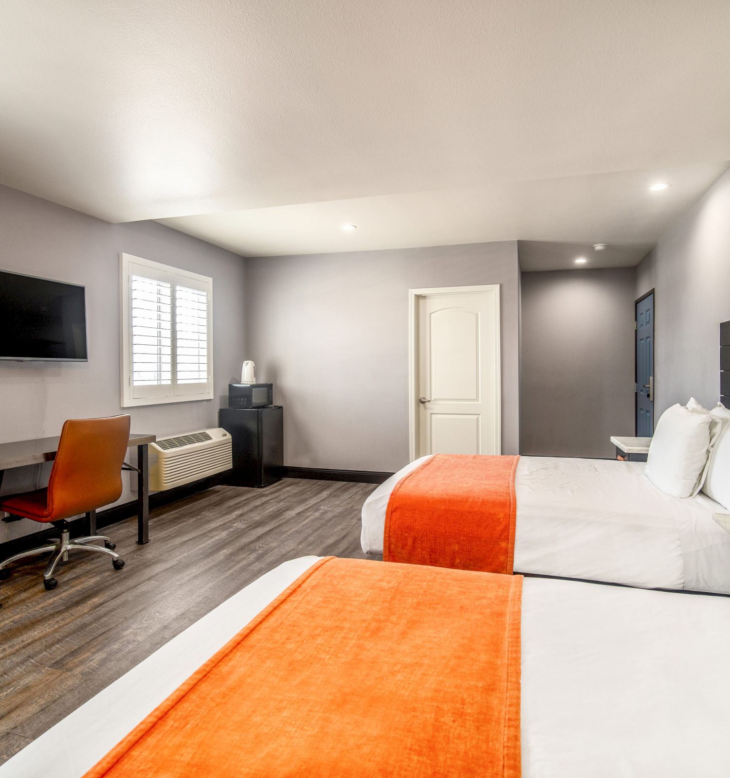 A modern hotel room features two beds with orange blankets, a wall-mounted TV, a desk with a chair, and a door leading to an adjoining space.