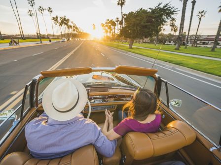 A couple is driving in a convertible at sunset on a palm-lined road, holding hands and enjoying the scenic view.