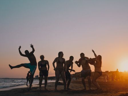 A group of six people is enjoying a beach at sunset, with one person playfully jumping while others strike fun poses against the setting sun.