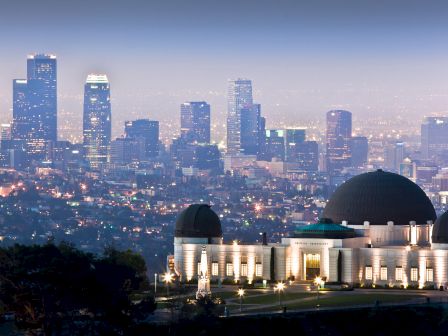 A domed building is foregrounded with a city skyline filled with skyscrapers in the background, all illuminated under evening light.