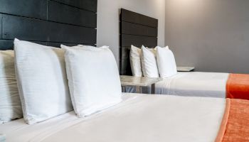 The image shows two neatly made beds with white linens and multiple pillows, featuring black headboards and accentuated by orange throws at the foot.