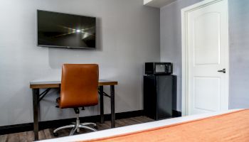 The image shows a room with a wall-mounted TV, a desk with a brown chair, a microwave, a mini-fridge, and a closed white door.