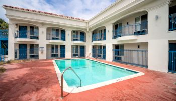 Outdoor swimming pool surrounded by a two-story motel building with balconies and blue railings, featuring a red concrete pool deck.