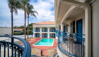 The image shows a two-story hotel or motel with a central outdoor swimming pool, palm trees, and blue doors with balconies overlooking the pool.