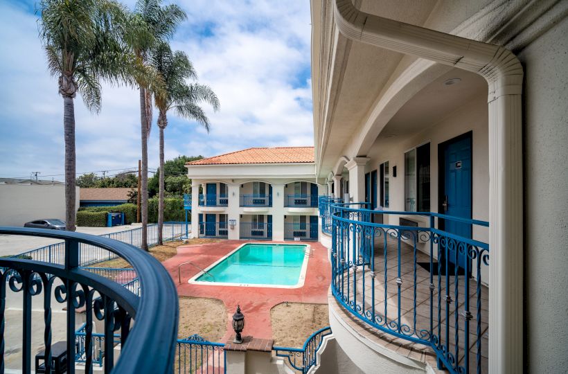 The image shows a two-story hotel or motel with a central outdoor swimming pool, palm trees, and blue doors with balconies overlooking the pool.