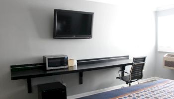 The image shows a room with a mounted TV, a microwave, a small refrigerator, a desk with a chair, and a window to the right.