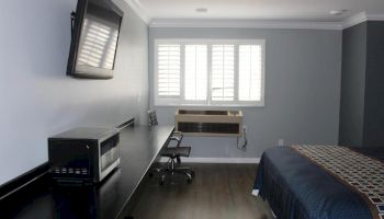 The image shows a modern bedroom with a desk, chair, microwave, and TV, plus a bed with a blue bedspread near a window with shutters.