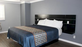 A neatly arranged bedroom with a large bed, blue bedding, and pillows against a dark headboard, in a room with gray walls and wooden flooring.