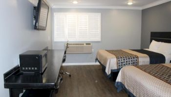 The image shows a tidy hotel room with two beds, a desk with a microwave, a wall-mounted TV, and a window with blinds, ending the sentence.