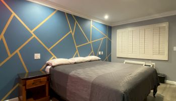 A bedroom with a geometric accent wall, a bed with gray bedding, side tables, and wooden flooring. The room has recessed lighting and a window.