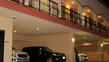 This image shows three cars parked under a building with balconies and ornate railings above them. The parking area is illuminated with lights.