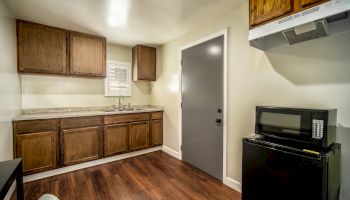 A small kitchen with wooden cabinets, a gray door, a black microwave on a mini-fridge, and wooden flooring.