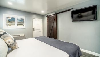 A bedroom with a neatly made bed, a wooden sliding door, a TV mounted on the wall, a white door, and a window with white shutters.