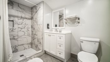 A modern bathroom with a marble-tiled shower, white vanity with a mirror, toilet, and towel rack, featuring gray floor tiles and minimal decor.