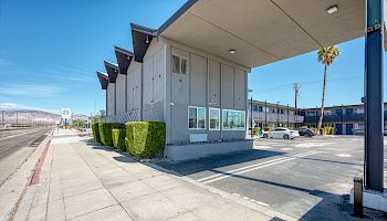 A modern, two-story, angular building with parking spaces and hedges, located near a deserted sidewalk under a clear blue sky.