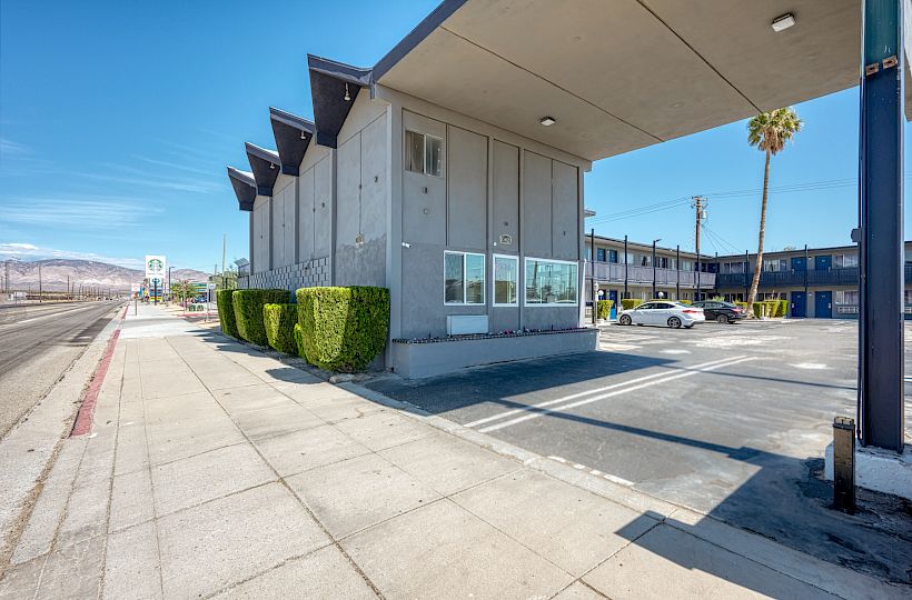 A modern, two-story, angular building with parking spaces and hedges, located near a deserted sidewalk under a clear blue sky.