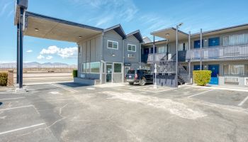 A two-story motel with exterior corridors, parking lot, and covered entry. Blue doors and railings, surrounded by a rural landscape, clear sky.