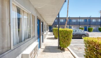 A motel exterior with a walkway, green bushes, parked cars, and windows under a clear blue sky.