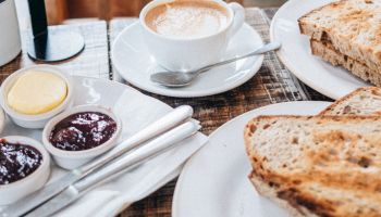 A cozy cafe setting with a cup of coffee, a plate of toast, and small bowls of jam and butter on a wooden table with a chair in the background.