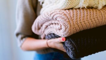 A person is holding a stack of folded, knitted blankets or sweaters in neutral colors, including white, beige, and gray.