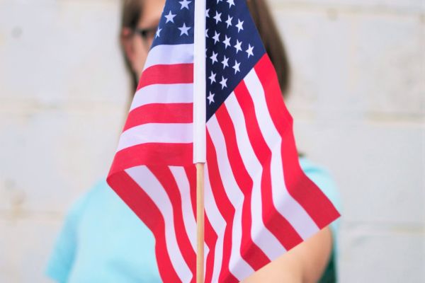 A person wearing a light blue shirt is holding a small U.S. flag in front of them, obscuring part of their face. The background is a white wall.