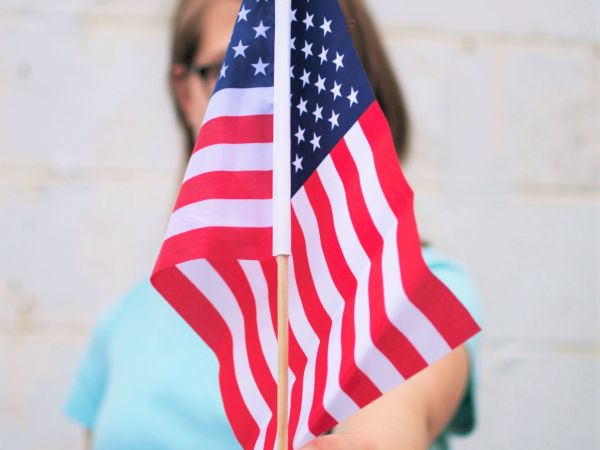 A person wearing a light blue shirt is holding a small U.S. flag in front of them, obscuring part of their face. The background is a white wall.