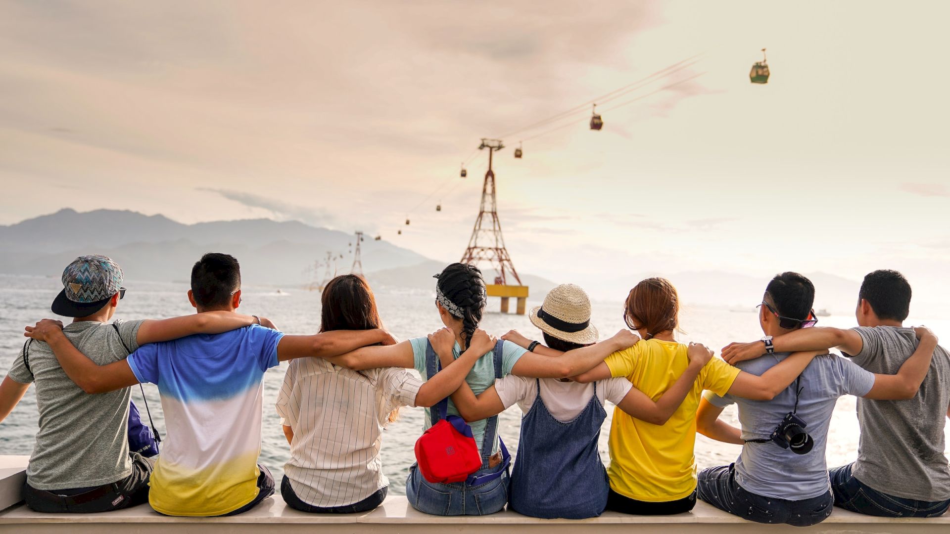 A group of seven people sit side by side with their arms around each other, facing a body of water with cable cars and mountains in the background.