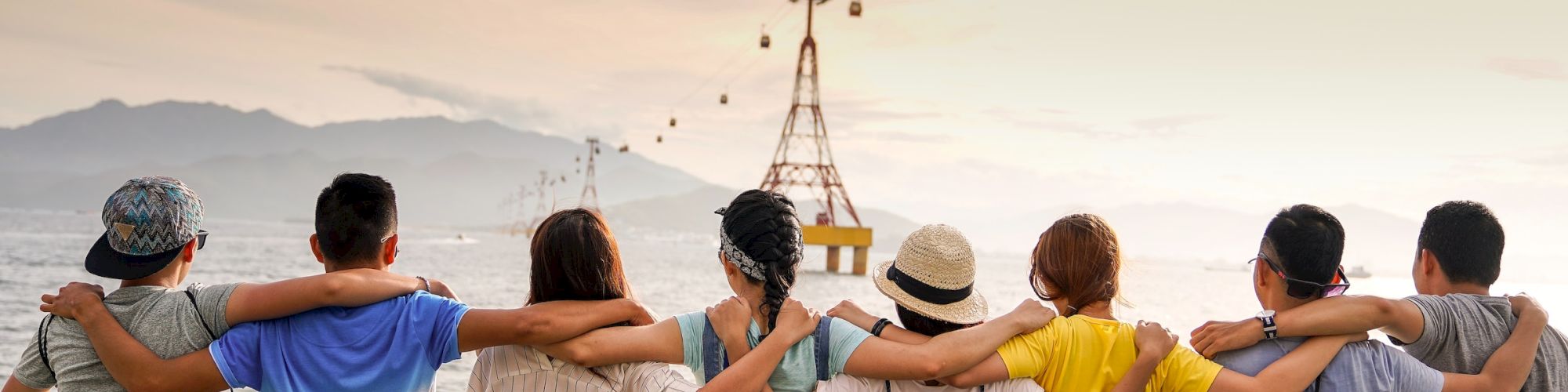 A group of seven people sit side by side with their arms around each other, facing a body of water with cable cars and mountains in the background.