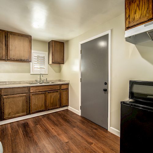 A small kitchen with wooden cabinets, a gray door, a microwave on a black mini-fridge, a sink, and a white range hood on a wood floor.