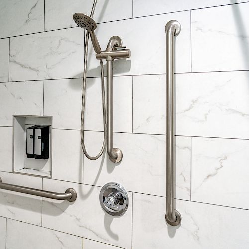 A modern shower with white tile wall featuring a handheld showerhead, safety grab bars, soap dispensers, and a control knob.