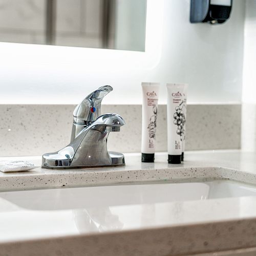 A bathroom sink with a chrome faucet, two tubes of toiletries, and two small packets lies on a light-colored countertop.