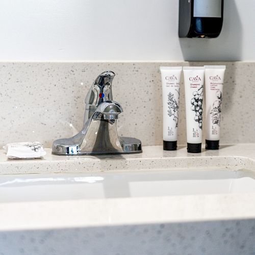 A bathroom countertop with a sink, a silver faucet, three cosmetic tubes, and some small packets placed near the faucet’s base.