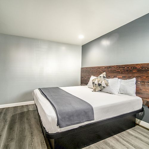 A modern hotel room features a bed with white linens, wooden headboard, gray walls, a wall-mounted TV, and wood-look flooring.