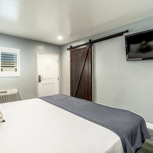 A modern bedroom with a neatly made bed, wooden sliding door, wall-mounted TV, gray walls, and a window.