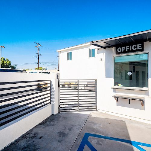 The image shows a small office building with a gated entry and a service window, under a clear blue sky, and a sign that says 