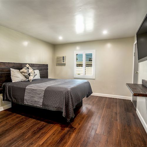 A modern bedroom with a large bed, wooden floors, a wall-mounted TV, and a small window with shutters.