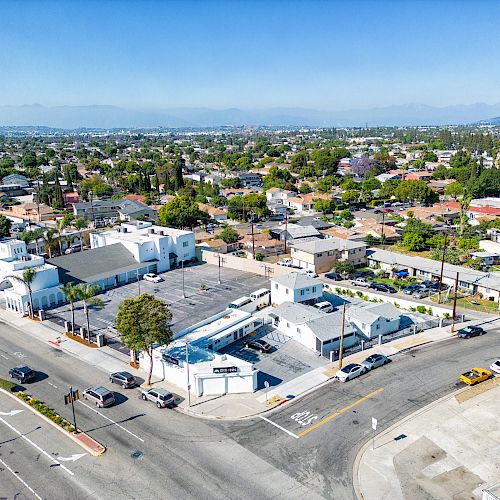 An aerial view of a suburban area featuring residential homes, streets, a church with a dome, and a parking lot on a clear day with mountains in the background.