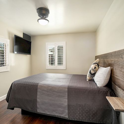 A small bedroom with a bed, a wooden headboard, a wall-mounted TV, two windows with shutters, and a ceiling light fan.