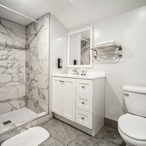 A modern bathroom featuring a marble-tiled shower, white vanity with a mirror, toilet, and towel rack on a neutral-toned wall.