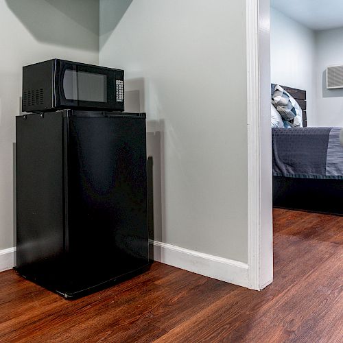 A compact black microwave sits on top of a mini refrigerator against a light-colored wall, near a doorway leading to a bedroom with a bed and an air conditioner.