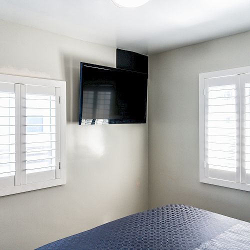 A bedroom corner with a wall-mounted TV, two windows with white shutters, and part of a bed with a blue cover is shown in the image.