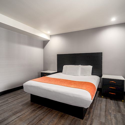 A modern, minimalist hotel room with a large bed featuring a white bedspread and an orange runner, nightstands, and grey walls.