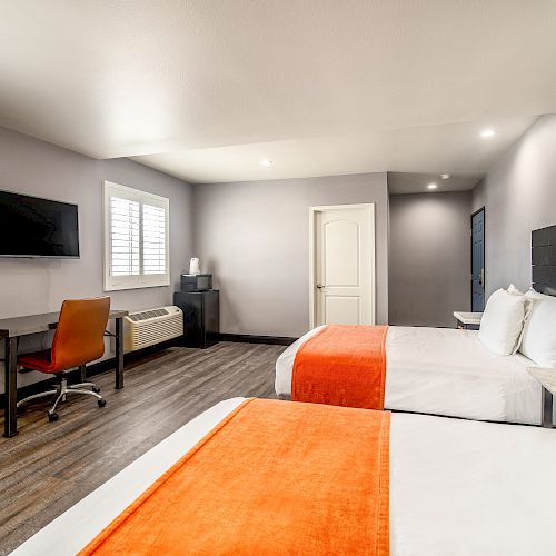 The image shows a modern hotel room with two beds, orange accents, a desk with a chair, a wall-mounted TV, and a window with blinds.