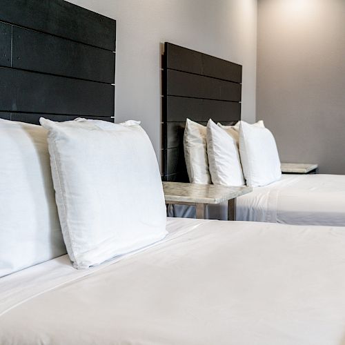 The image shows two neatly made beds with white linens and four pillows each, against a gray wall with dark wood headboards, and orange throws at the foot.