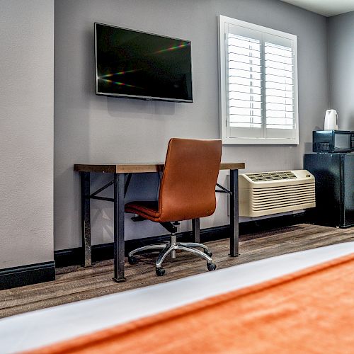 The image shows a hotel room with a desk, chair, wall-mounted TV, window with blinds, air conditioner, and a small fridge with a white appliance on top.