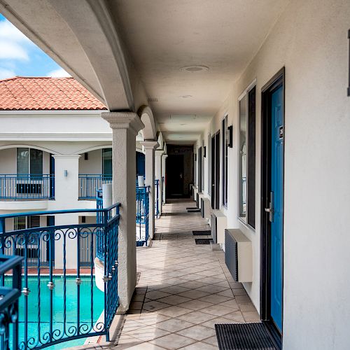 A hotel corridor with blue doors and railings overlooks a pool area, featuring archways and tiled flooring in a Mediterranean-style design.