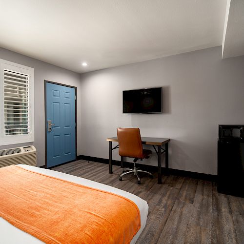 A modern hotel room with a bed, desk, TV, blue door, window, and air conditioner; has wood flooring and minimalist decor.